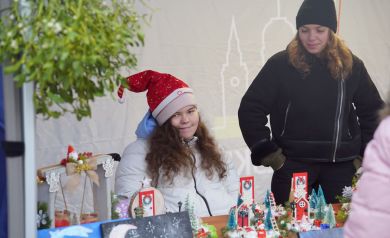 Na zdjęciu dwie dziewczyny. Jedna ubrana jest na czarno, druga ma założoną na głowie czapkę Mikołaja. Przed nimi widać stroiki świąteczne. 