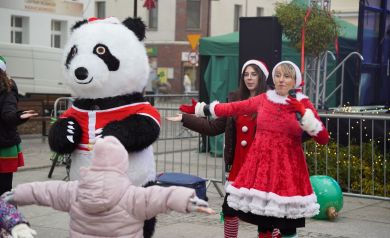 Animacje z dużą maskotką pandy. Na zdjęciu widać dwie kobiety w świątecznych strojach i dziecko w różowej czapce i różowej kurtce.