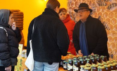 Rozmowa mężczyzny w kapeluszu z kobietą w czarnej bluzie. Widać dwie inne kobiety również. Na stołach znajdują się słoiki z miodami. 