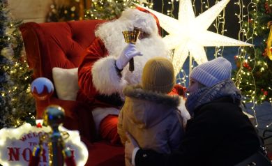 Kobieta z dzieckiem obok mężczyzny przebranego za Mikołaja. Mężczyzna w dłoni ma dzwonek. 