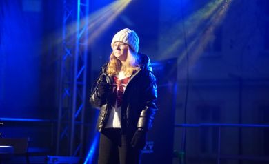 Na zdjęciu dziewczyna stojąca na scenie trzyma mikrofon dłoni. 