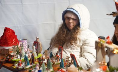 Na zdjęciu dziewczynka o kręconych włosach z czapką i kapturem na głowie. Przed nią ręcznie wykonane ozdoby świąteczne. 