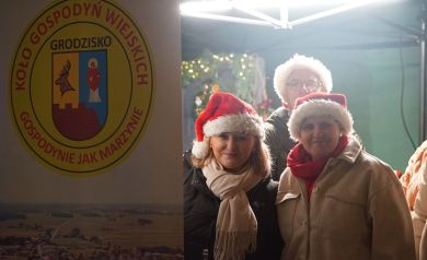 Trzy kobiety w świątecznych czapkach przy banerze Koła Gospodyń Wiejskich z Grodziska. 
