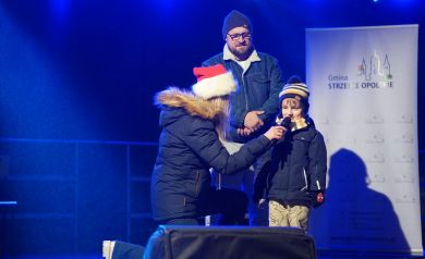 Na oświetlonej scenie dziecko w zimowej kurtce i czapce mówi do mikrofonu trzymanego przez kobietę w mikołajkowej czapce, a obok stoi dorosły mężczyzna. W tle widać baner gminy i sceniczne oświetlenie.