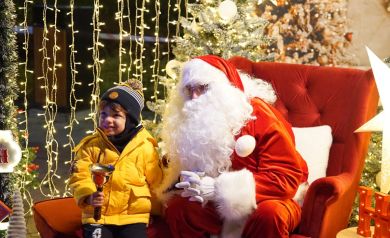 Chłopiec w żółtej kurtce trzyma dzwonek w dłoni i dzieci obok mężczyzny przebranego za Mikołaja, który siedzi na czerwonym fotelu. Zdjęcie wykonane jest w namiocie przystrojonym świątecznymi dekoracjami. 