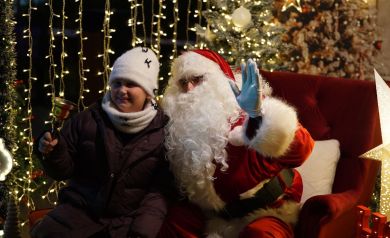 Dziewczynka trzymająca dzwonek w dłoni, obok niej mężczyzna w stroju Mikołaja. Zdjęcie wykonane jest w namiocie świątecznie udekorowanym. 