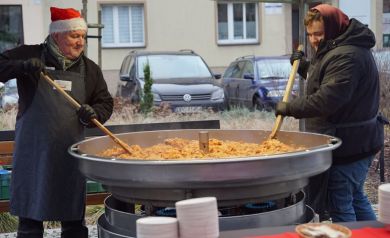 Dwóch mężczyzn miesza dużymi łyżkami drewnianymi bigos w dużej patelni. 