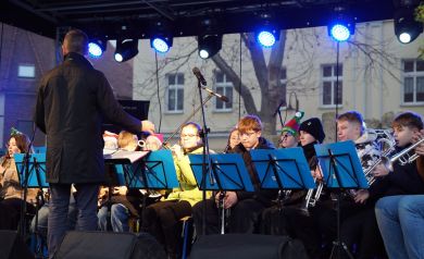 Występ orkiestry dętej. Przed muzykami stoi dyrygent i pulpity z niebieskimi teczkami. 