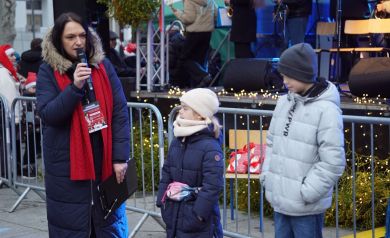 Kobieta mówi do mikrofonu. Obok niej stoi dziewczynka i chłopiec. W tle widać scenę. 