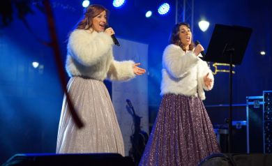 Kobiety w błyszczących sukniach i futrach śpiewają do mikrofonów. 