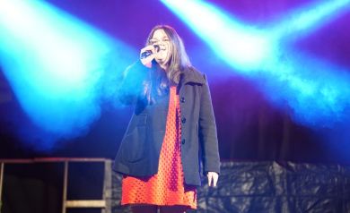 Dziewczyna w czerwonej sukience i płaszczu stoi na scenie i śpiewa do mikrofonu. 