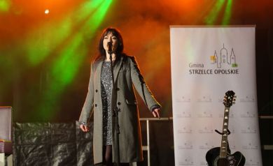 Kobieta w błyszczącej sukience i płaszczu śpiewa do mikrofonu. Obok niej stoi gitara i baner gminy Strzelce Opolskie. 