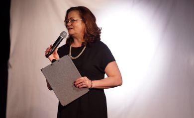 Kobieta w okularach, w czarnej sukience i koralach mówi do mikrofonu trzymanego w prawej dłoni. W lewej dłoni trzyma szarą teczkę. 