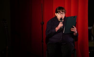 Kobieta w czarnych okularach w prawej dłoni trzyma mikrofon, do którego mówi, zaś w lewej dłoni ma teczkę. 