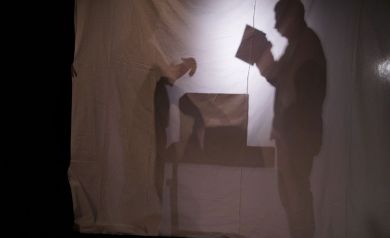 Zdjęcie przedstawia teatr cieni, na którym widoczne są sylwetki dwóch osób oraz mebel, prawdopodobnie stół.
