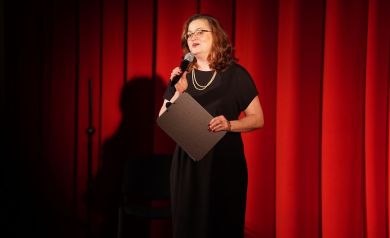 Na zdjęciu kobieta w czarnej sukience i koralach, przemawia do mikrofonu. W jednej dłoni trzyma szarą teczkę. Za kobietą rozciąga się czerwona kotara 