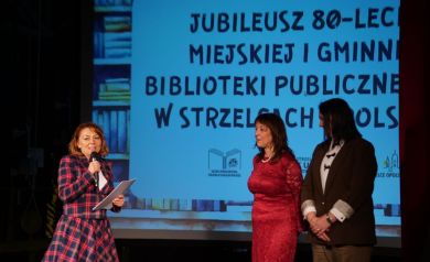 Na zdjęci trzy kobiety. Kobieta ubrana w marynarkę i spódnicę w kratkę przemawia do mikrofonu. W tle widać wyświetlaną prezentację. 