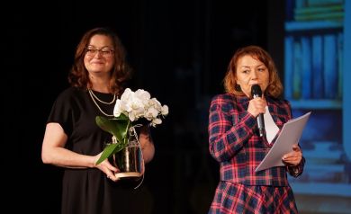 Na zdjęciu dwie kobiety. Jedna ubrana w czarną sukienkę trzyma w dłoni kwiat w doniczce. Druga ubrana w marynarkę i spódnicę w kratkę przemawia do mikrofonu, trzyma w dłoni również białe kartki. 