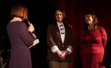 Na zdjęciu trzy kobiety. Jedna ubrana jest w bordową sukienkę, druga w białą koszulę z czarnymi kokardami, brązową marynarkę i brązowe spodnie, a trzecia w czerwoną, koronkową sukienkę. Kobieta ubrana w bordową sukienkę przemawia do mikrofonu. 