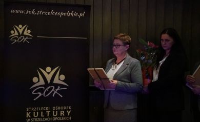 Na zdjęciu dwie kobiety, jedna trzyma w dłoni ramkę, druga kwiat w doniczce. Obok nich stoi czarny baner Strzeleckiego Ośrodka Kultury. 