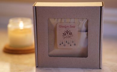 Na zdjęciu znajduje się małe, brązowe, kartonowe pudełko prezentowe z przezroczystym okienkiem. Na naklejce znajdującej się na pudełku napisane jest ,,Wesołych Świąt ŻYCZY SOK'.