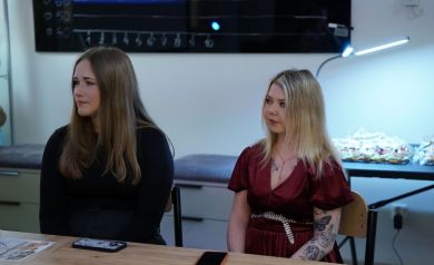 Na zdjęciu dwie kobiety jedna ubrana w bordową sukienkę, druga w czarny sweter. Siedzą przy stoliku.