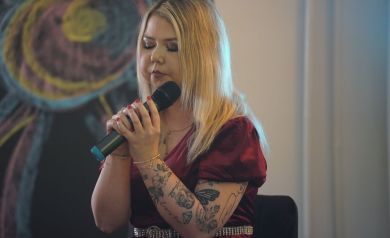 Na zdjęciu młoda kobieta w blond włosach podczas swojego występu. Ubrana jest w bordowa sukienkę.