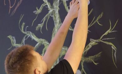 Młody mężczyzna mocuje znak graficzny na czarnej tablicy z rysunkiem drzewa i napisem ,,Drzewo Marzeń'.