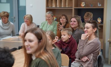 Na zdjęciu dorośli i dzieci siedzący przy stolikach. W tle widać regały drewniane.