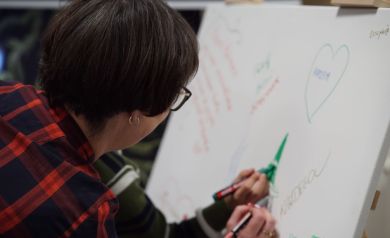 Kobieta w krótkich włosach, okularach i czerwonej sukience w kratkę pisze markerem po płótnie. Obok niej w tle widać dziecko, również malujące markerem po płótnie.