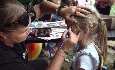 Na zdjęciu widać kobietę malującą dziecku twarz. Kobieta trzyma pędzelek w jednej ręce i delikatnie podtrzymuje głowę dziewczynki drugą ręką, aby precyzyjnie wykonać malunek. Dziewczynka siedzi na krześle, ma jasne włosy spięte w kucyk z różową spinką. W tle widać stół z różnymi materiałami do malowania twarzy oraz inne dzieci i dorosłych.