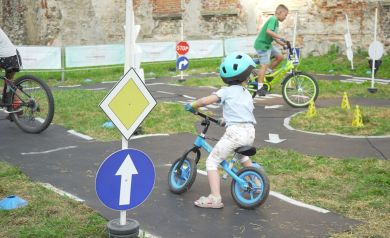 Na zdjęciu widać dzieci jeżdżące na rowerach po miasteczku ruchu drogowego, z oznakowanymi uliczkami i znakami drogowymi. Najmłodsze dziecko jedzie na rowerku biegowym w kasku ochronnym. Całość odbywa się w bezpiecznym, edukacyjnym otoczeniu – to atrakcja przygotowana w ramach wydarzenia „Zakręcona Wakacyjna Meta”.