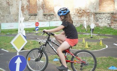 Zdjęcie pokazuje jedną z atrakcji ,,Zakręconej Wakacyjnej Mety', jaką jest Rowerowa Akademia. Dziewczynka jeździ na rowerze po miasteczku rowerowym. Trasa z oznakowaniem drogowym uczy zasad ruchu w bezpieczny i zabawowy sposób.