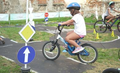 Na zdjęciu widać dziewczynkę jadącą na rowerze po specjalnie przygotowanym miasteczku ruchu drogowego. Ma na sobie kask ochronny, a wokół niej ustawione są znaki drogowe. Na drugim palnie widać inną dziewczynkę, również jeżdżąca na rowerze oraz widać ruiny zamku. To edukacyjna atrakcja w ramach wydarzenia „Zakręcona Wakacyjna Meta”, ucząca dzieci zasad bezpiecznego poruszania się po drogach.