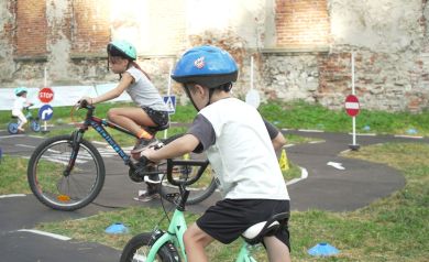 Na zdjęciu widać trójkę dzieci jeżdżących na rowerach na specjalnym placu do nauki jazdy. Wszystkie dzieci mają na głowach kaski ochronne. Plac jest oznakowany różnymi znakami drogowymi, takimi jak stop czy zakaz wjazduy. W tle widać starą, częściowo zniszczoną ścianę ruin zamku oraz trawę. Jest jasno, co wskazuje na porę dnia - prawdopodobnie jest to popołudnie.