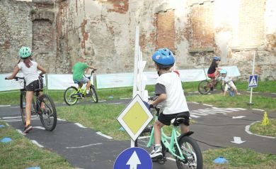 Na zdjęciu widzimy grupę dzieci na rowerach, które jeżdżą po wyznaczonym torze rowerowym. Tor jest oznaczony znakami drogowymi, takimi jak znak pierwszeństwa przejazdu i znak nakazu jazdy prosto. Dzieci mają na głowach kaski. Otoczenie to miejsce na świeżym powietrzu z trawą i częściowo zniszczonymi, starymi murami zamku w tle. 