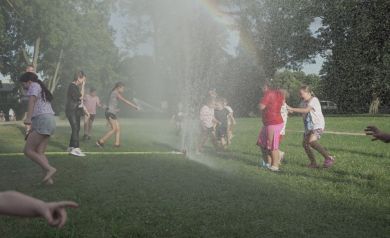 Na zdjęciu widać grupę dzieci bawiących się na trawie w parku lub na łące. Dzieci stoją wokół rozpryskującej się wody z węża. Woda tworzy delikatną mgiełkę, a w promieniach słońca widoczna jest tęcza. Niektóre dzieci są mokre, inne starają się unikać wody, ale wyglądają na zadowolone i radosne. W tle widać drzewa oraz fragmenty otoczenia parku.