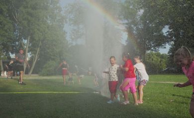 Na zdjęciu widzimy grupę dzieci bawiących się na trawie w parku. Dzieci są ochlapywane wodą ze spryskiwaczy, które tworzą mgiełkę wodną. W powietrzu widać tęczę. Dzieci wyglądają na bardzo zadowolone i radosne, bawią się w upalny dzień. W tle widoczne są drzewa oraz kilku dorosłych. 
