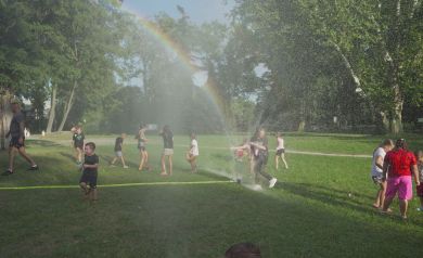 Na zdjęciu widać grupę dzieci bawiących się na świeżym powietrzu na trawniku. Dzieci są ochlapywane przez wodę ze spryskiwaczy, co tworzy przyjemną mgiełkę wodną. W tle, na niebie, pojawia się tęcza. Otoczenie to park z drzewami i zieloną trawą. 