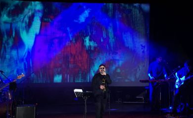 Na scenie śpiewający aktor podczas spektaklu muzycznego. W tle ekspresyjna grafika performatywna.