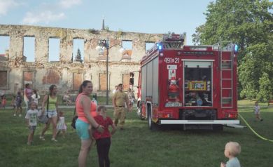 Na zdjęciu widać wóz strażacki, który był jedną z atrakcji wydarzenia „Zakręcona Wakacyjna Meta”. Dzieci i dorośli z zainteresowaniem podchodzą do pojazdu, a strażacy chętnie prezentują jego wyposażenie. Całość odbywa się na tle ruin zamku, w otoczeniu zieleni i w radosnej atmosferze rodzinnego pikniku.