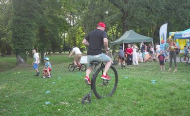 Na zdjęciu widać mężczyznę jadącego na oryginalnym, starym rowerze typu penny-farthing, który ma bardzo duże przednie koło i dużo mniejsze tylne. Mężczyzna ma na sobie czarną koszulkę, jasne spodenki i czerwone buty oraz czerwoną czapkę. W tle widać grupę dzieci i dorosłych. Są tam namioty, banery i dmuchane konstrukcje. 