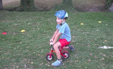 Na zdjęciu widzimy chłopca w niebieskim kasku i czerwonych spodenkach, który siedzi na bardzo małym, czerwonym rowerku. Chłopiec stoi na trawie, a w tle widać drzewa i oznaczenia z kolorowych pachołków na ziemi. 