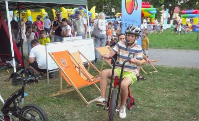 Na zdjęciu widzimy chłopca w kasku, jadącego na czerwonym rowerze. W tle jest plenerowa impreza z dmuchanymi zamkami, ludźmi i leżakami. 