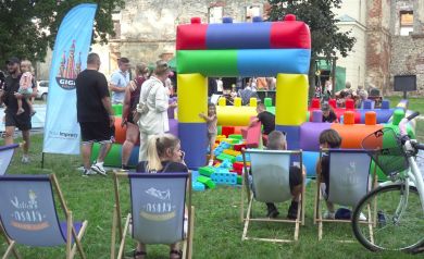 Na zdjęciu widać plenerowy festyn z dużą, kolorową dmuchaną konstrukcją do zabawy dla dzieci. Wokół bawią się dzieci i stoją dorośli, a na pierwszym planie kilka osób siedzi na leżakach. Tło stanowi park i stara zabytkowa ściana zamku.