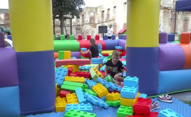 Na zdjęciu widać jedną z atrakcji wydarzenia „Zakręcona Wakacyjna Meta” – kolorową strefę zabaw z gigantycznymi klockami XXL. Dzieci bawią się w dmuchanym placu wypełnionym dużymi, klockami w różnych kolorach i kształtach. Dookoła widać fragmenty zrujnowanego zamku. Zabawa odbywa się na świeżym powietrzu, a dzieci z zaangażowaniem budują i eksplorują przestrzeń z klocków.
