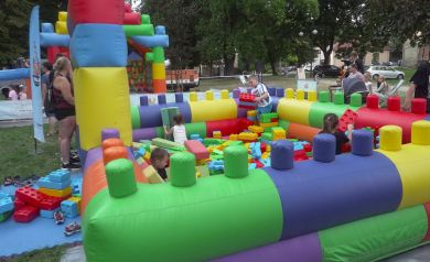 Na zdjęciu widać jedną z atrakcji jaką są gigantyczne klocki XXL. Wewnątrz bawią się dzieci, układając kolorowe, duże klocki. Wokół placu stoją dorośli i obserwują zabawę. Całość znajduje się na trawie, w parku.