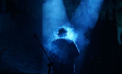 Scena ze spektaklu. Snopy światła spowite gęstą mgłą, po środku postać grającego aktora.