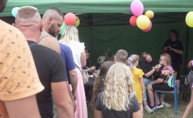 Na zdjęciu widzimy kolejkę ludzi ustawionych przed namiotem ozdobionymi kolorowymi balonami. W namiocie dwie osoby malują twarze dzieciom, które siedzą na krzesłach. Jedna dziewczynka ma już pomalowaną twarz na wzór psa. W kolejce czekają zarówno dorośli, jak i dzieci.