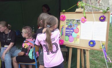 Na zdjęciu dzieci i młodzież oglądają tablicę z ogłoszeniami o wydarzeniu „Zakręcona Wakacyjna Meta”. W tle widać zielony namiot, osoby malujące twarze i osoby z pomalowanymi twarzami. 